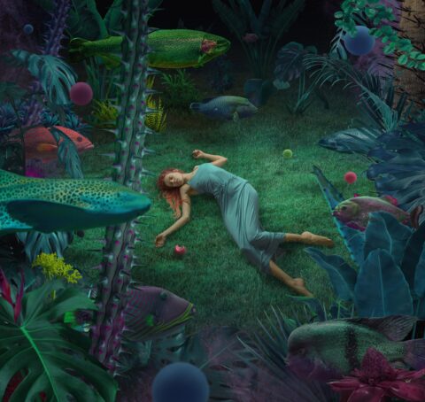 Woman with red hair and green dress lying on grass underwater with fish swimming by