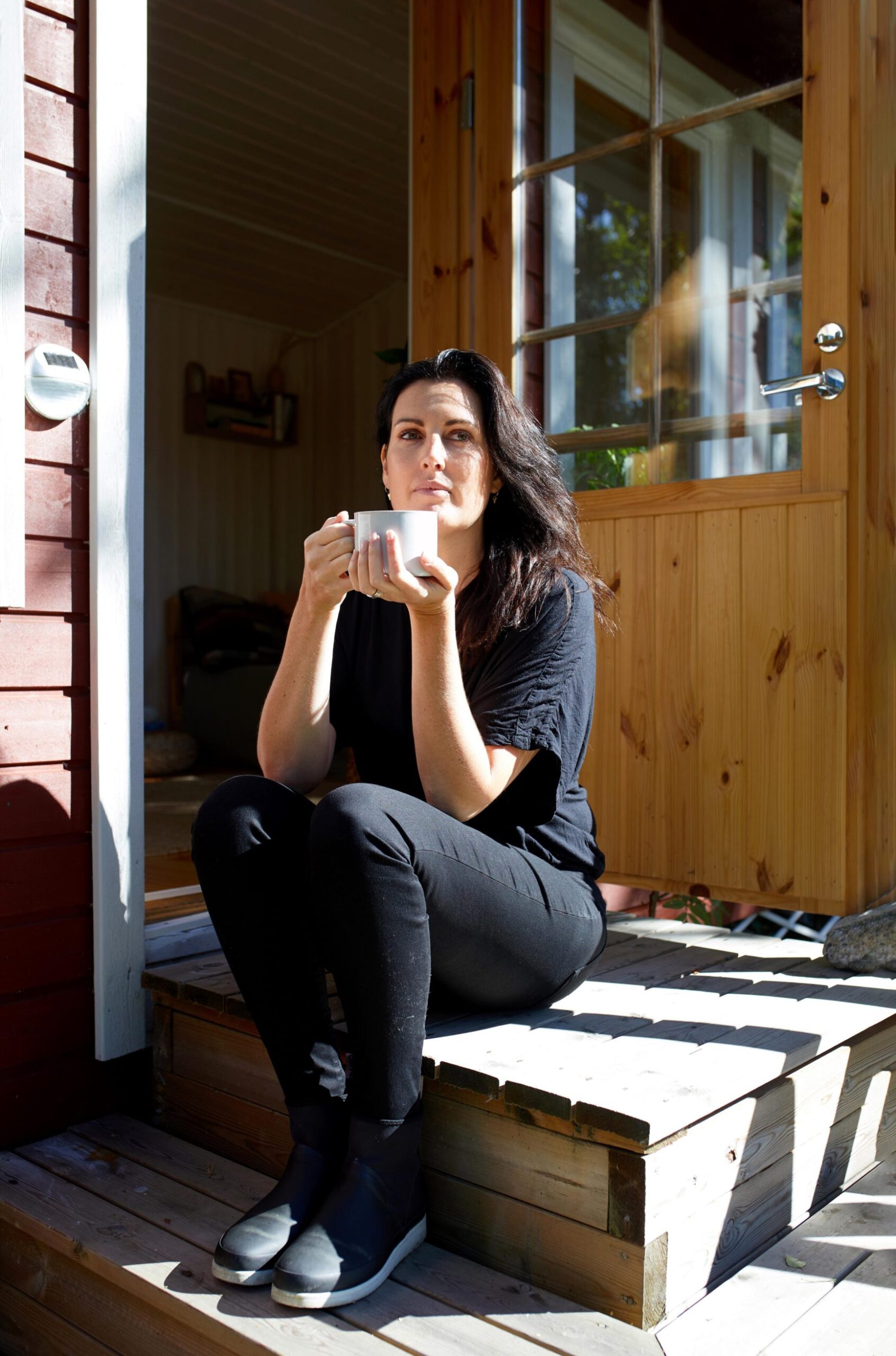 Melanie Dower sitting on the steps of her wooden cabin with a cup of tea