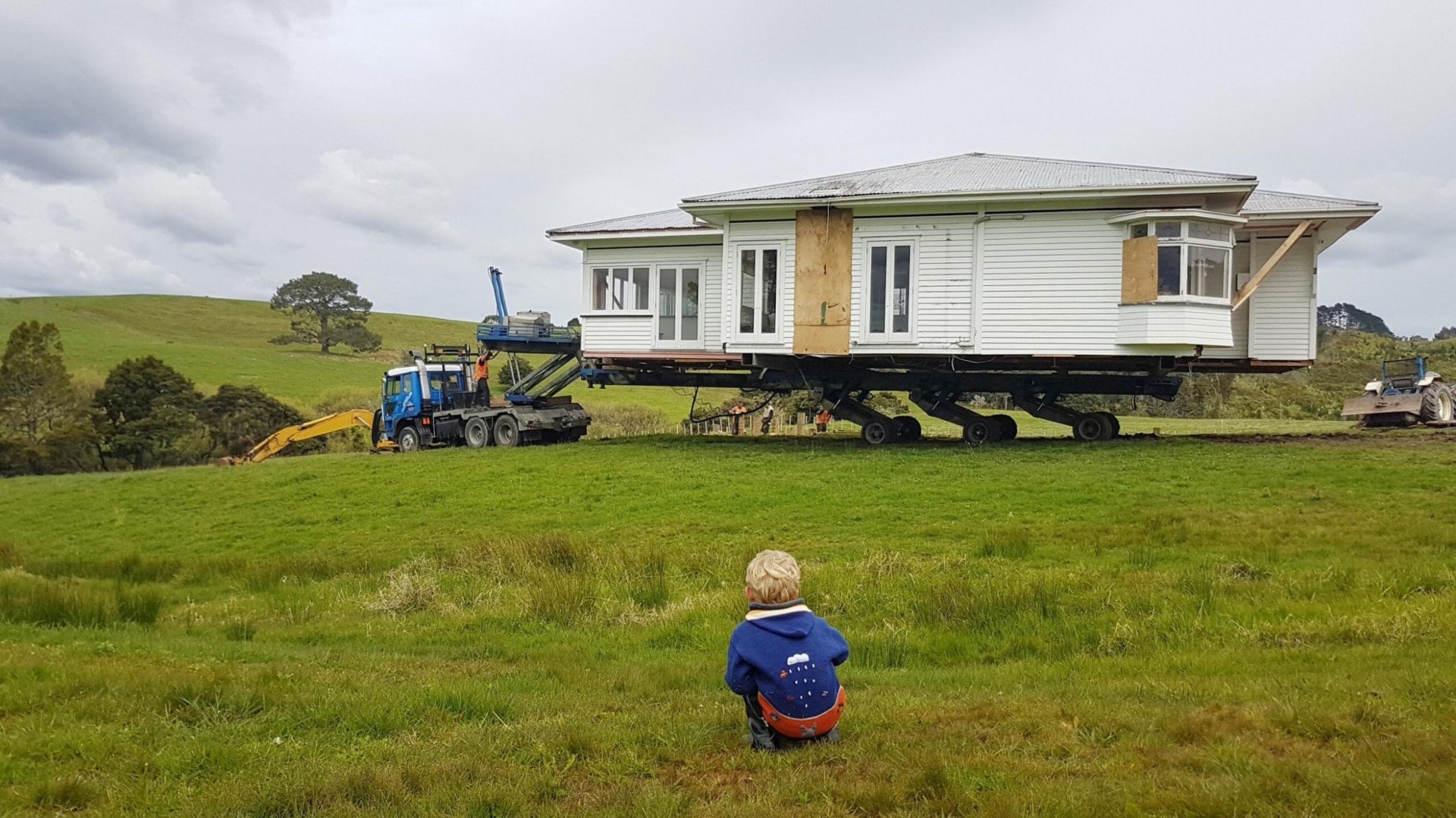 An old single story villa styled home with an angled roof being relocated by a tractor in a green field