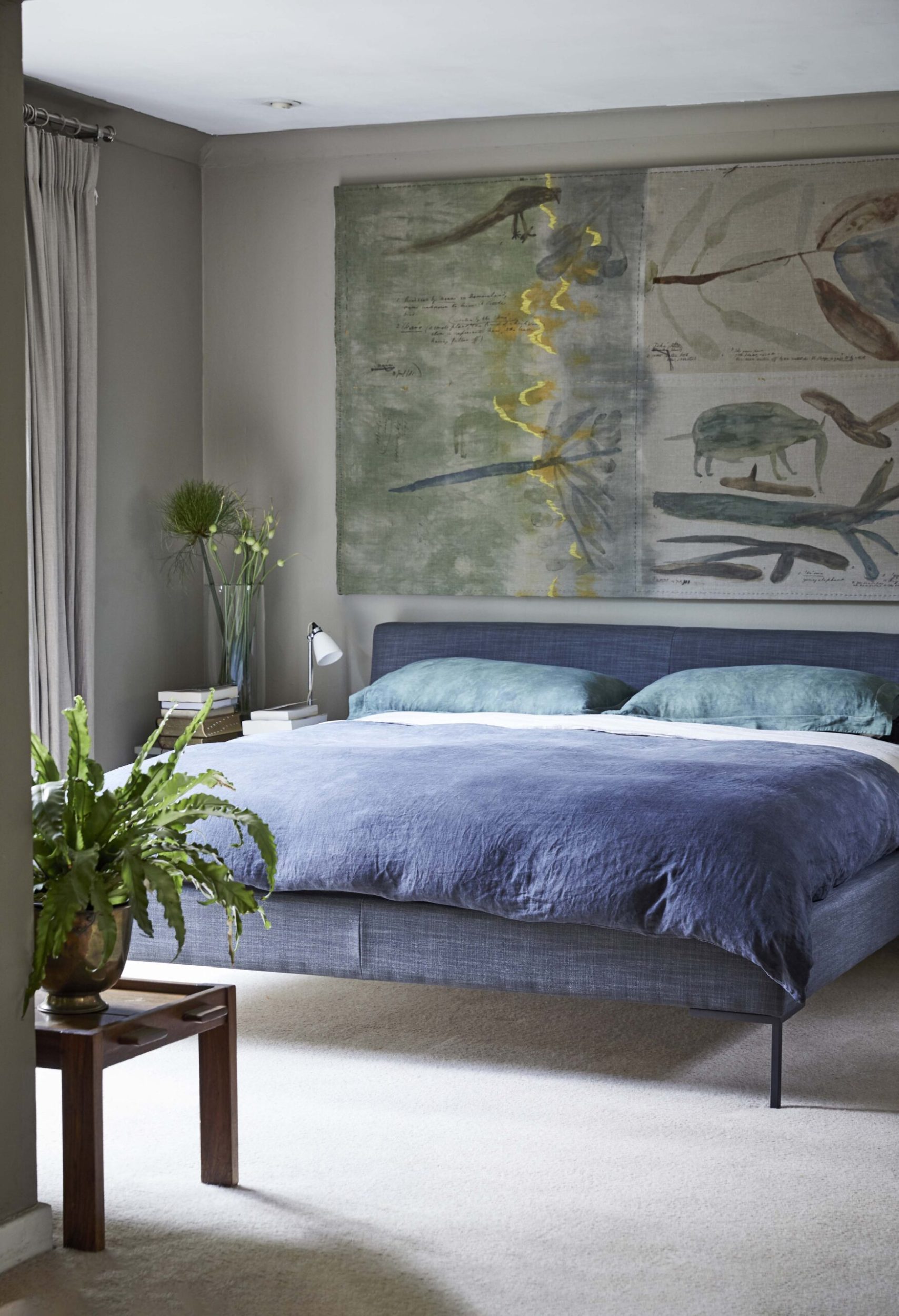 Bedroom with a grey wall, a white carpet, a large bed with a blue bedspread and a large statement artwork hanging above bed