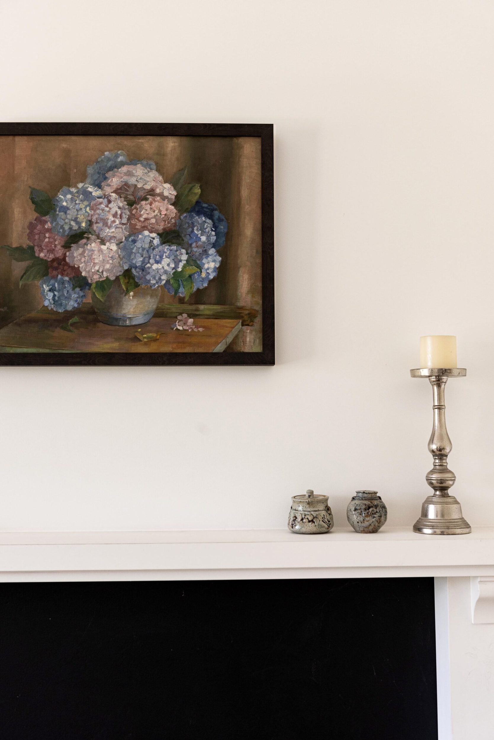 A white wall with a black fireplace, a floral hanging artwork and silver vintage candle holder