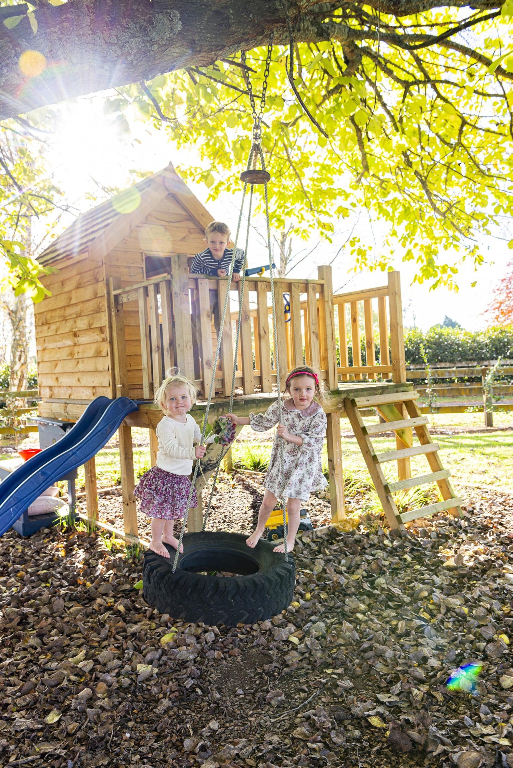 Two young girls playing on a tyre swing while a young boy looks out from a wood playhouse