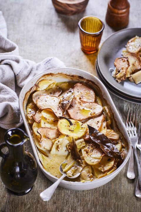 Potato, leek and pear bake served with cutlery on an oval shaped white dish set on top fo a brown table