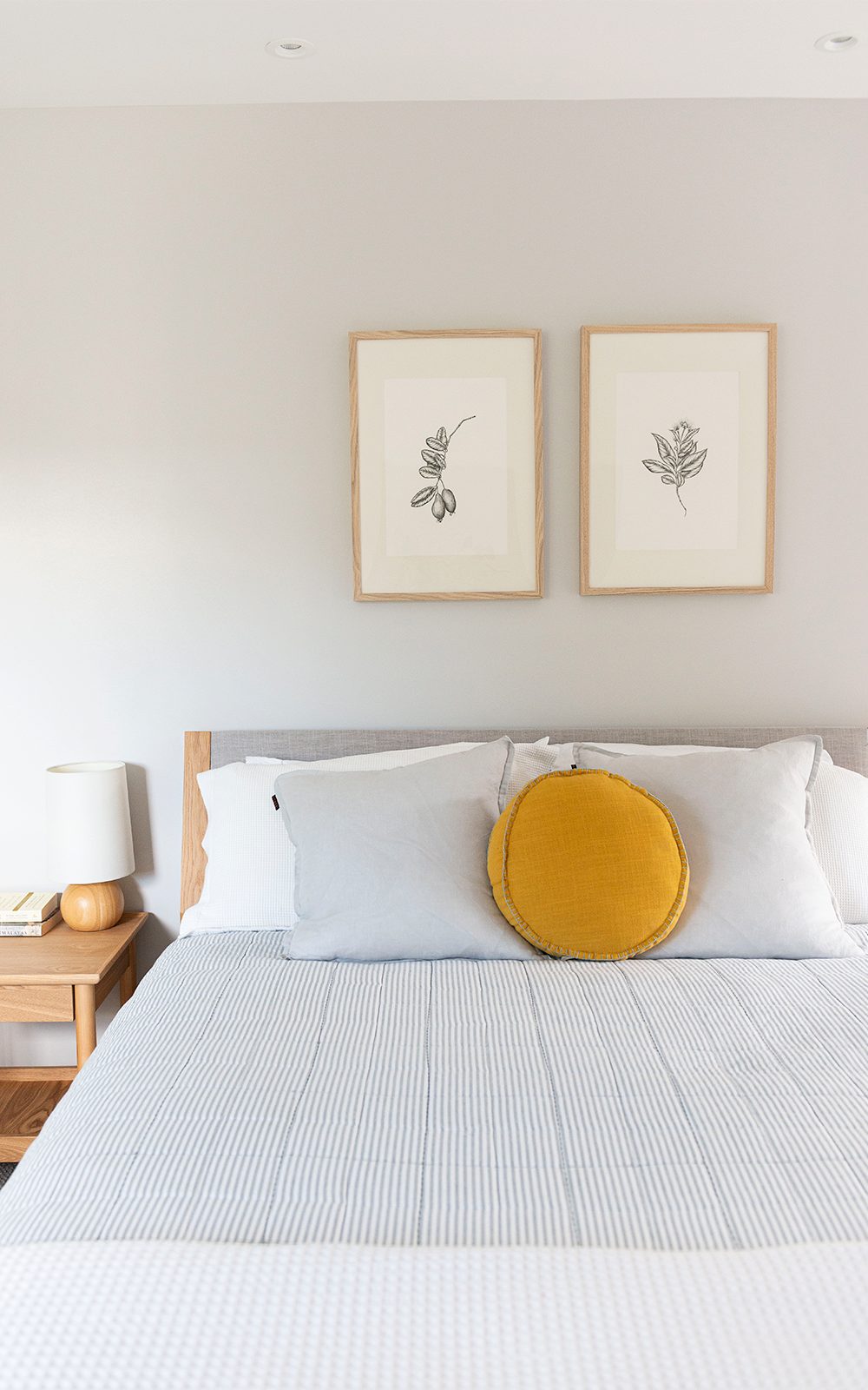 Bed with grey bedlinen, mustard coloured round pillow and two wooden framed artwork on the walls
