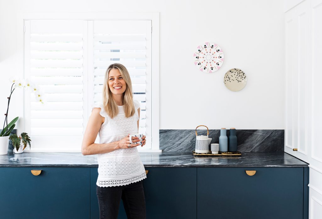 Hannah Gordon  smiles in front of kitchen bench with black marble and dark green cupboards