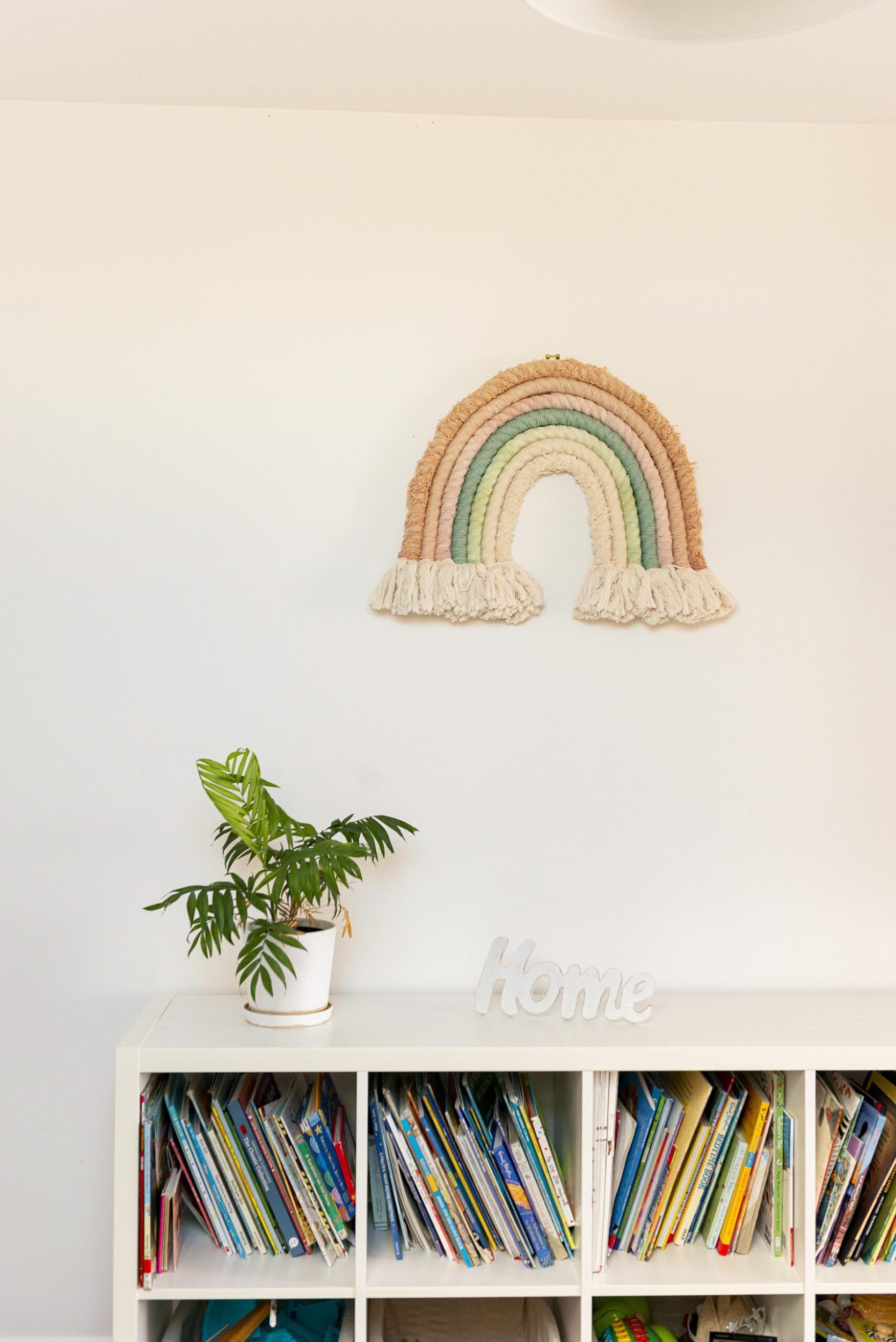 A rainbow macrame wall hanging on a white wall and a bookshelf on the floor below