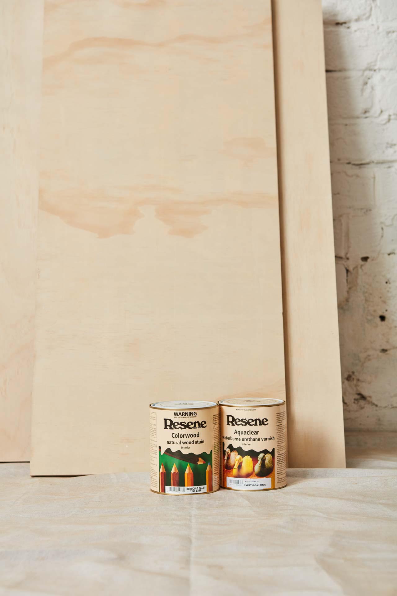 Two sheets of plywood leaning against the wall with buckets of Resene wood stain on the floor