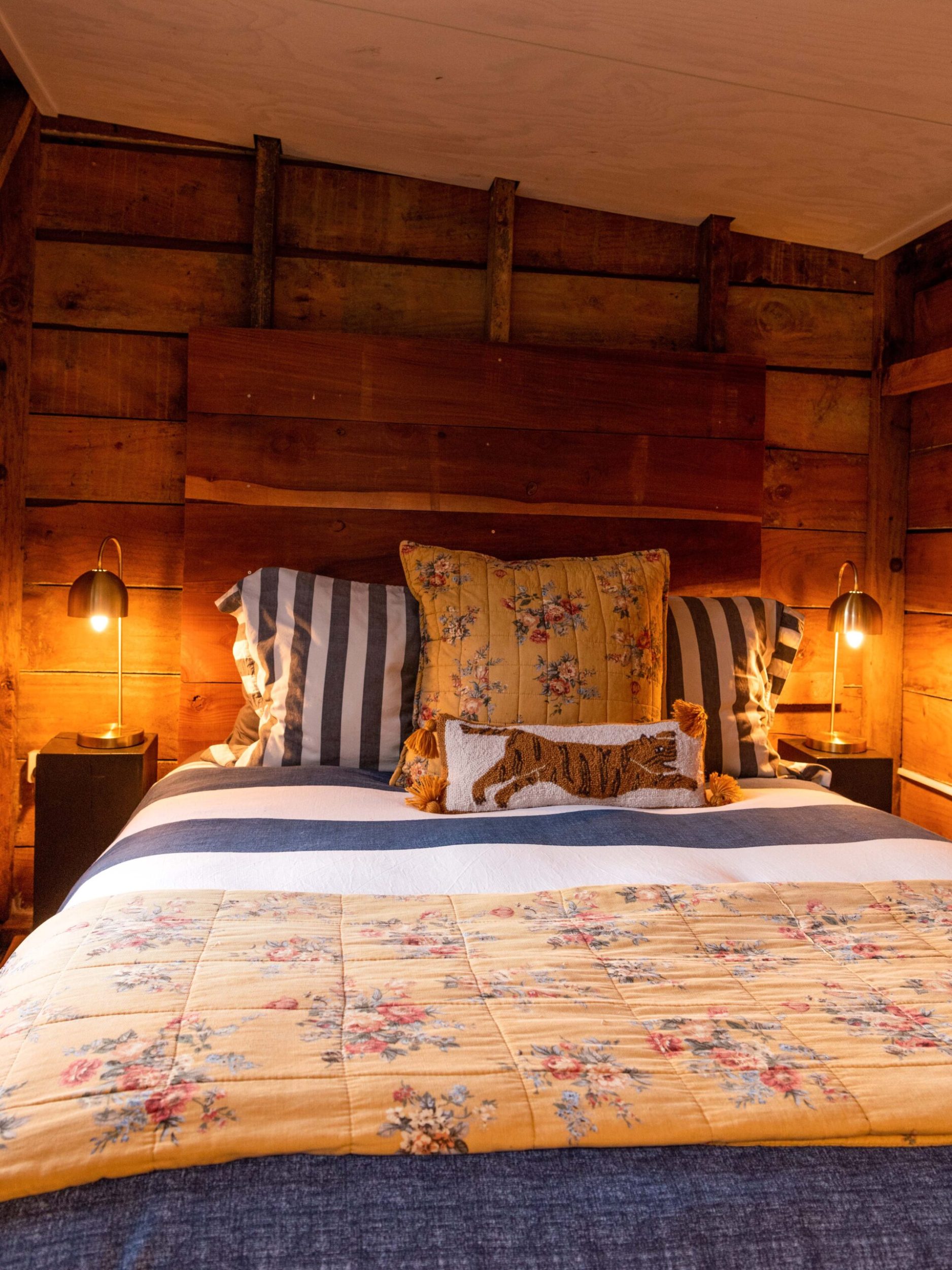A wood-panelled bedroom with a floral duvet, two lamps on either side of the bed and a cushion with a tiger on it