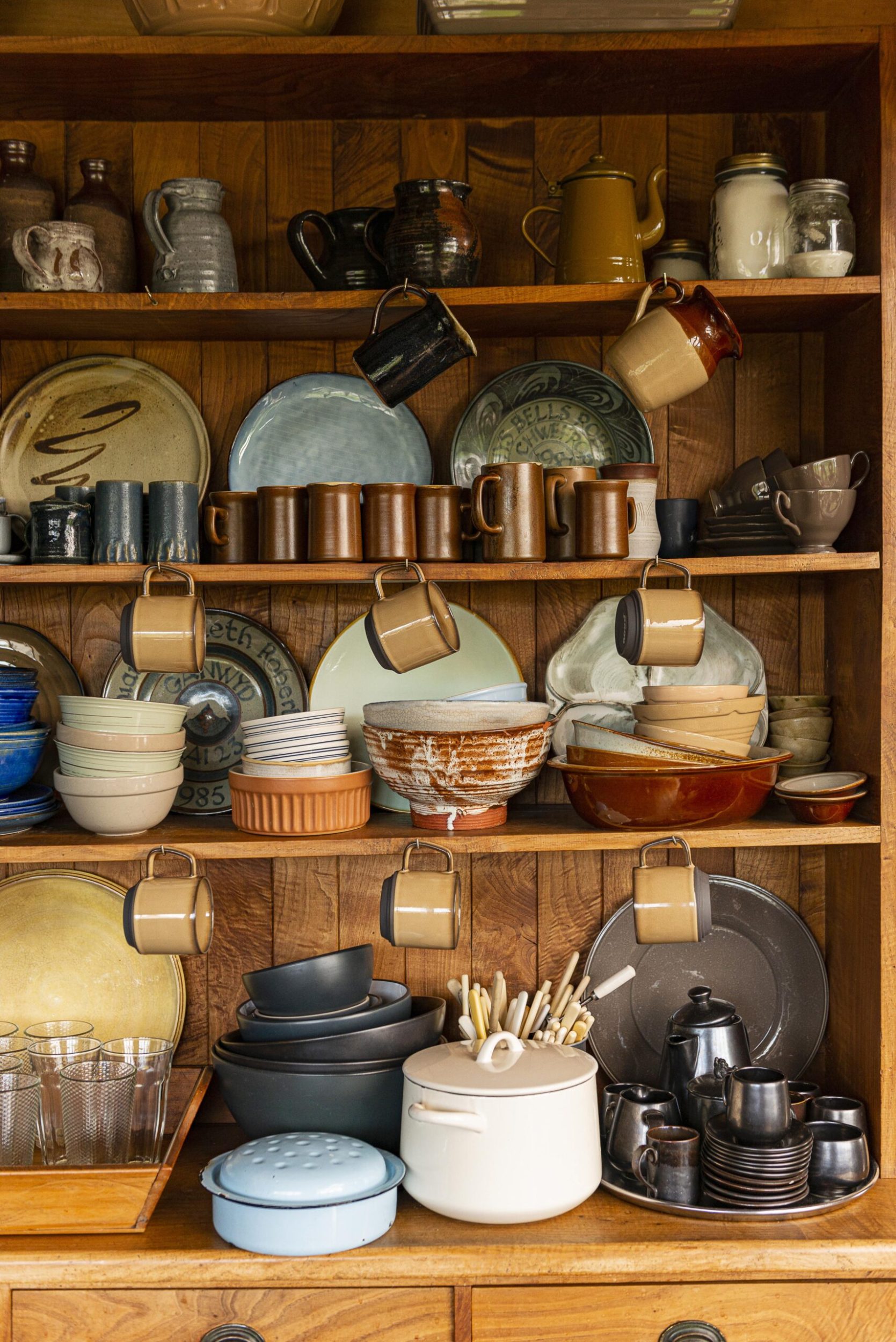 A wood shelving unit with shelves holding assorted bowls, hanging cups and plates