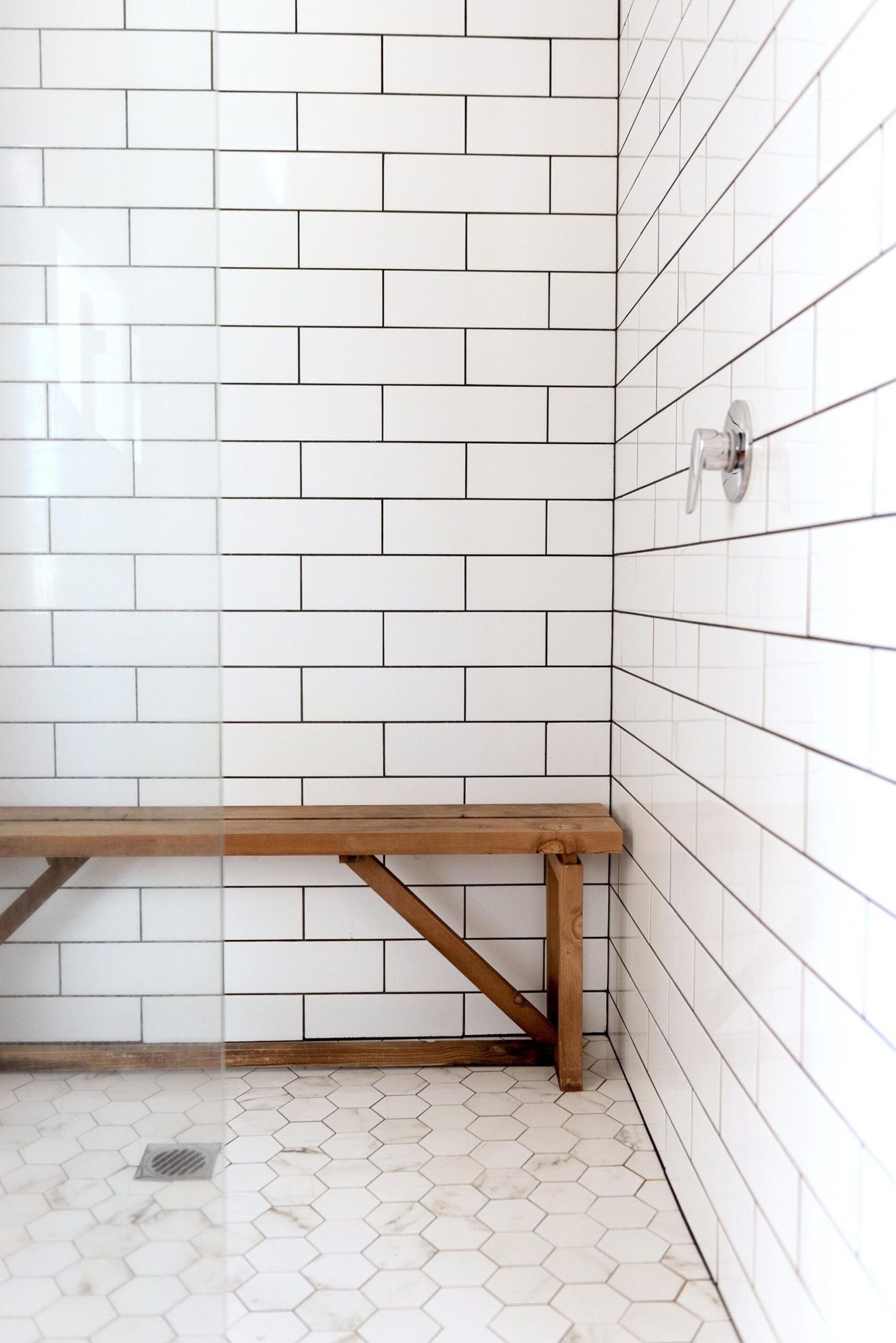 A large shower with a glass divider, white subway tiles, hexagonal floor tiles and a timber bench