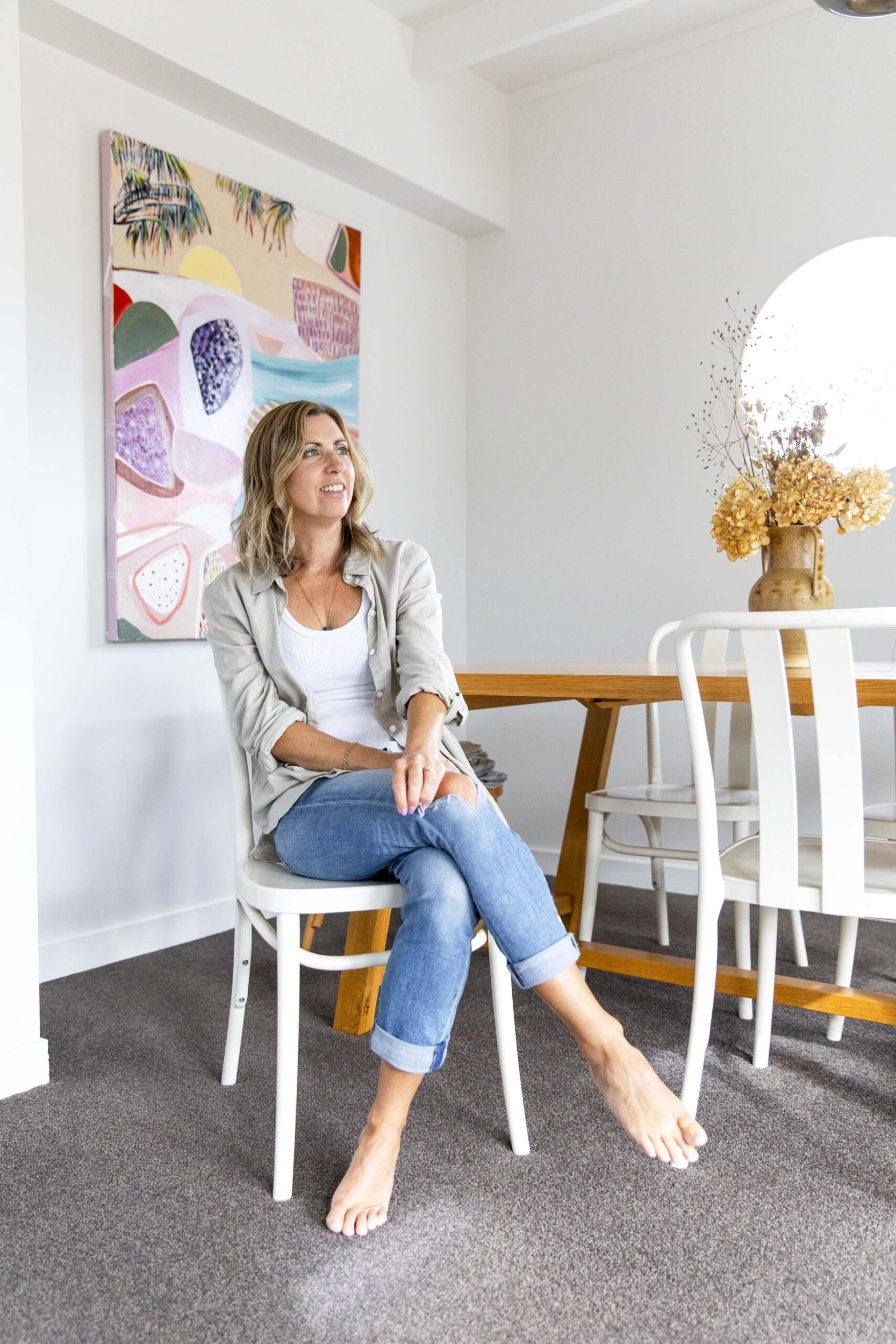 Brenda Clews sitting in her dining area with one of her artworks hung on wall behind her 