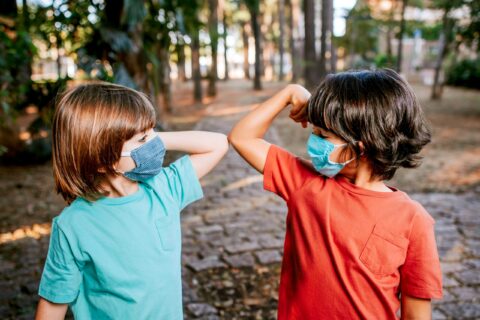 Child in blue t-shirt and child in red t-shirt wearing masks and touching elbows