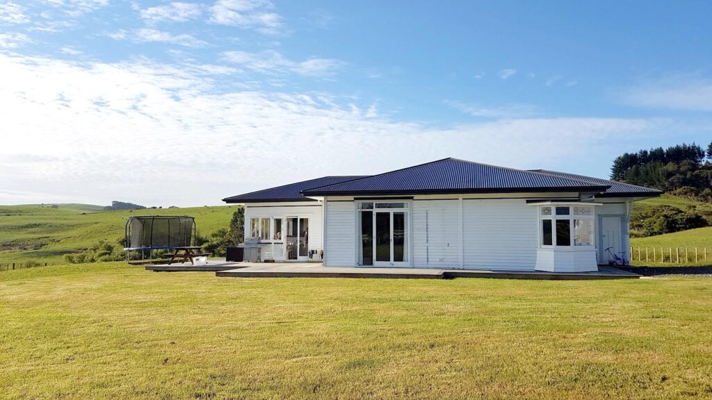 A single story home wth white cladding and a black roof in a green farm