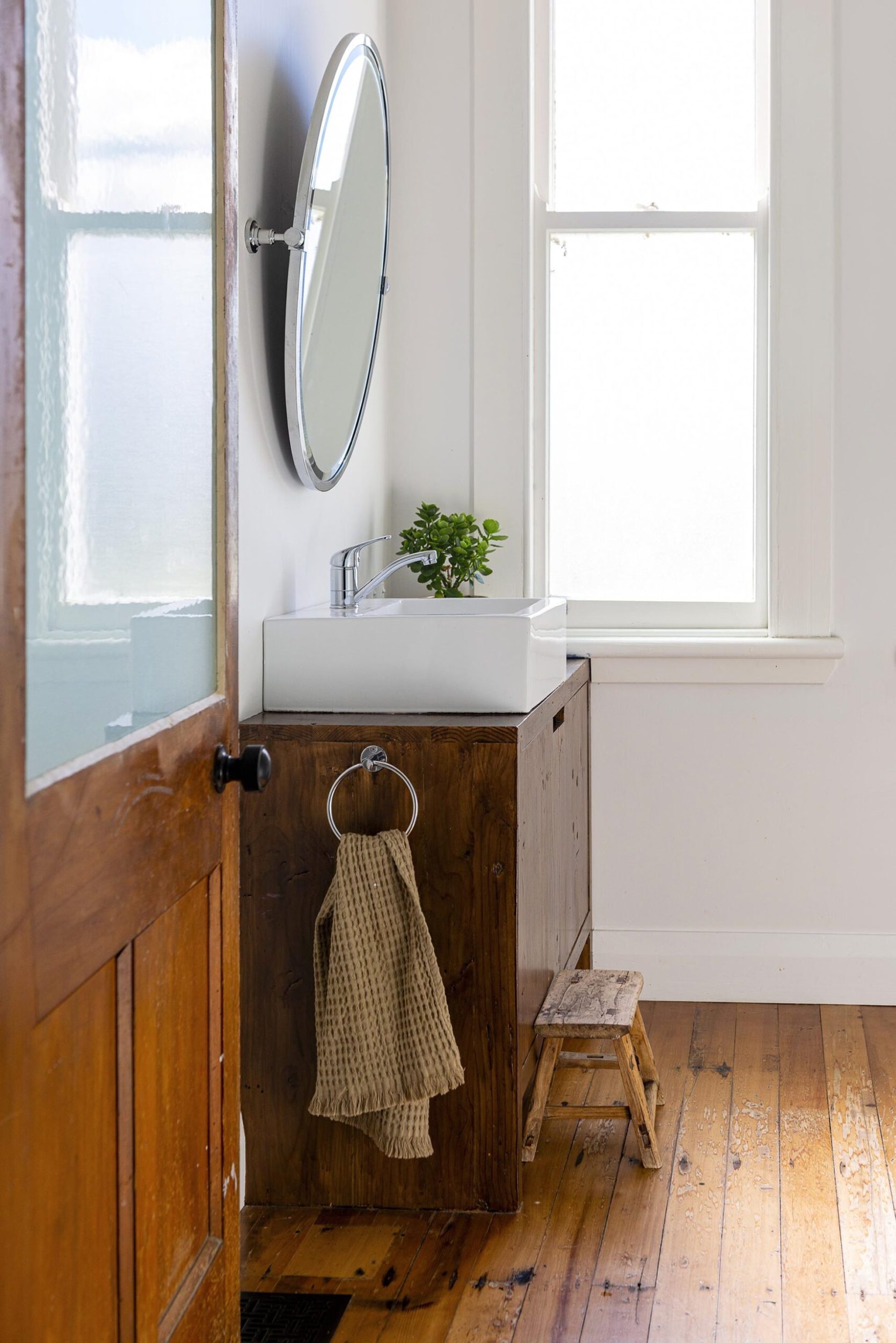 A bathroom with white walls, wooden floors, a round mirror and a wood sink cabinet 
