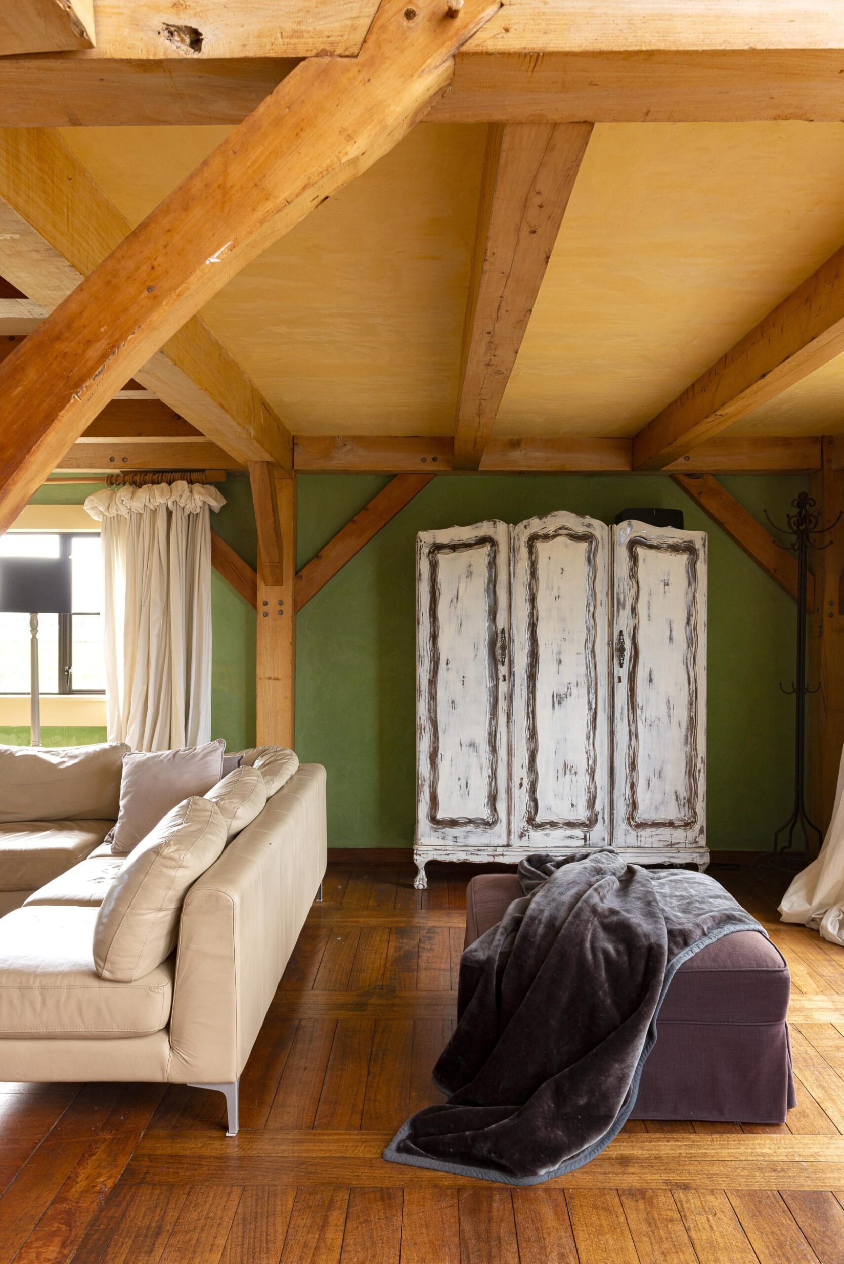 A living room area in a cottage with limewashed green walls, polished wood floors and a vintage whitewashed standing armoire