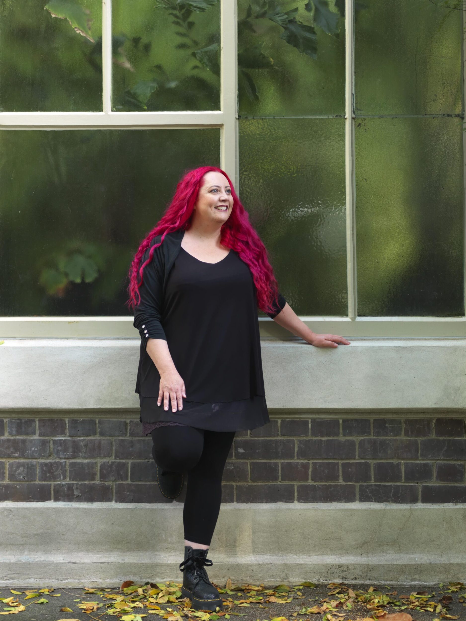 Siouxsie Wiles wearing a black top, cardigan and Doc Martens standing by a tall window