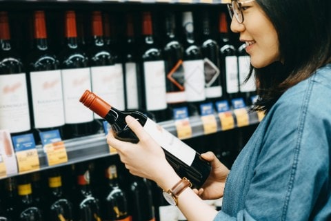 Young woman choosing and looking at bottle of wine in wine store