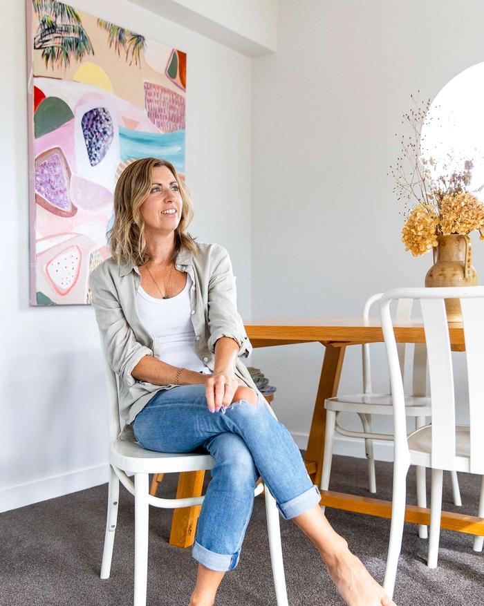 Brenda Clews sitting in her dining area with one of her artworks hung on wall behind her 