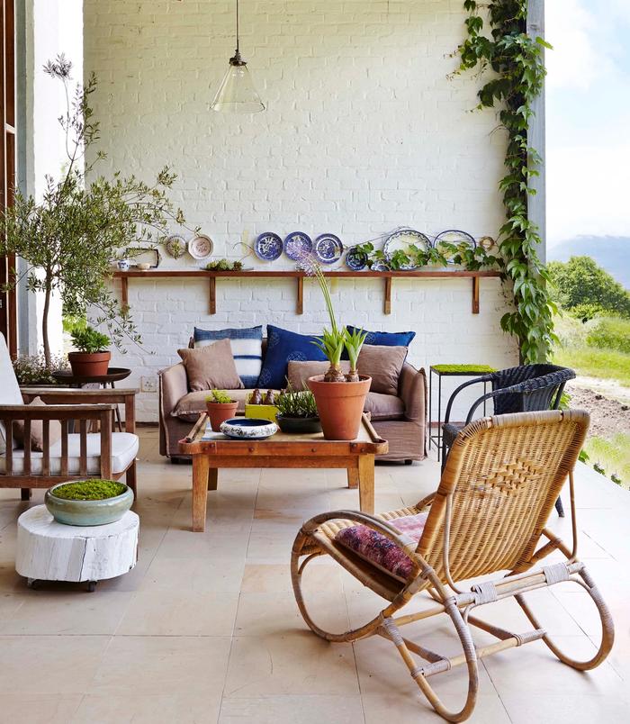 Country style outdoor room, with white painted brick wall, a floating shelf with hanging plates above it, small seating area with lounge chair and arm chair and a wicker rocking chair