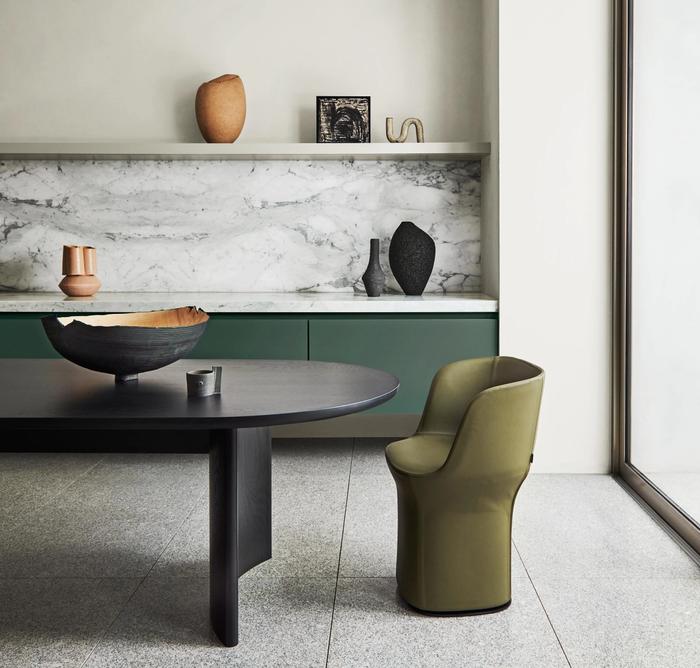 A dining room containing a curved wooden table, a green chair and a marble shelf with sculptures on it.