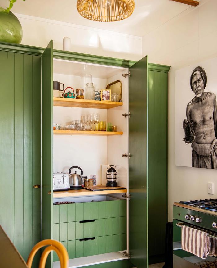 Green kitchen with pantry doors open to see open shelving and a bench top inside the cupboard