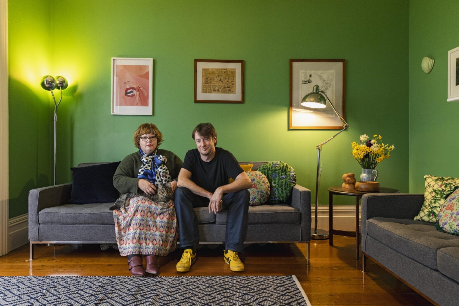 Faith Walmsley and husband Nicholas Cole sit on a grey couch in front of a green wall