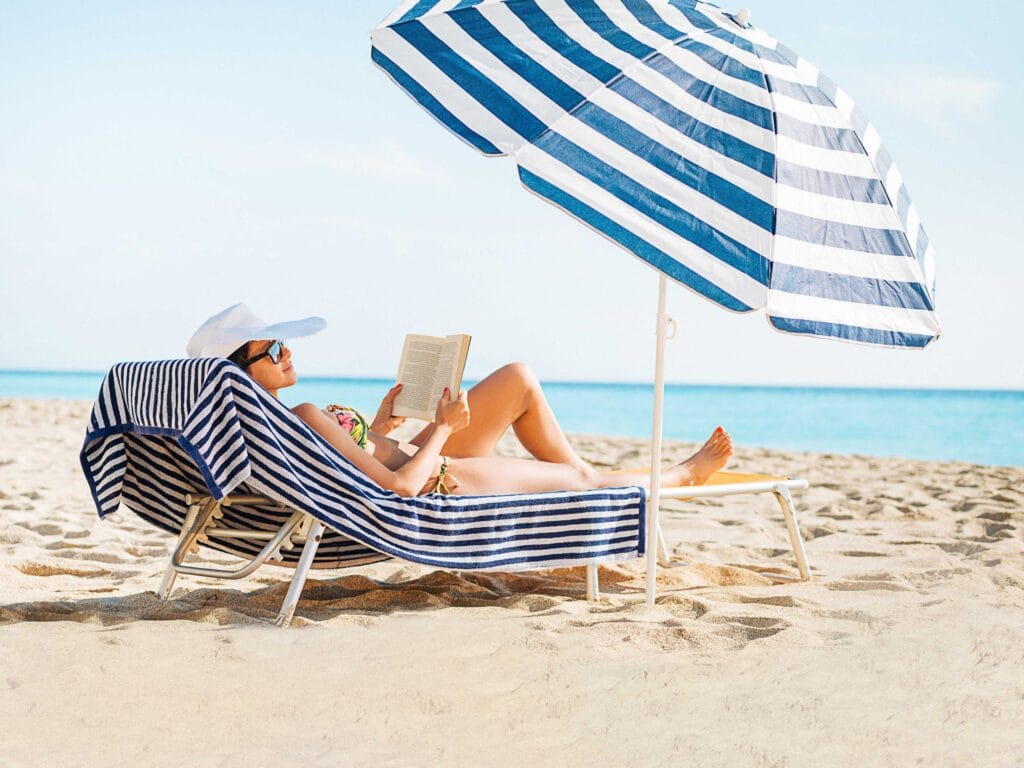 Woman reading on the beach under a blue striped umbrella on sun lounger