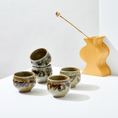 Five vintage ceramic mugs on a table with a wavy wooden vase in the background