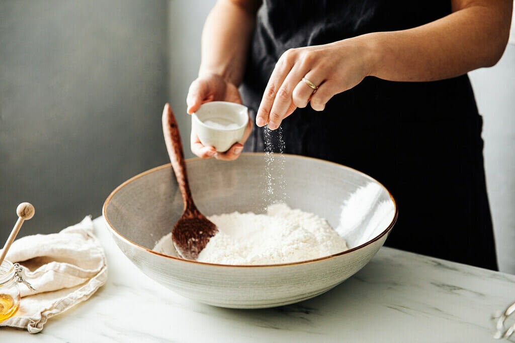 Women pinching salt into a bowl of flour. Bowl sitting on white marble table alongside honey in a jar