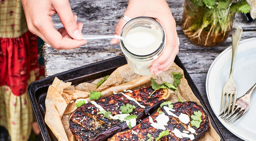 Person holding jar of sauce spooning it over eggplant on tray