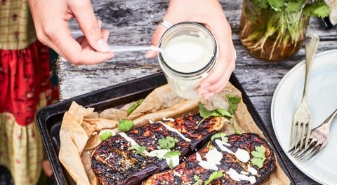 Person holding jar of sauce spooning it over eggplant on tray