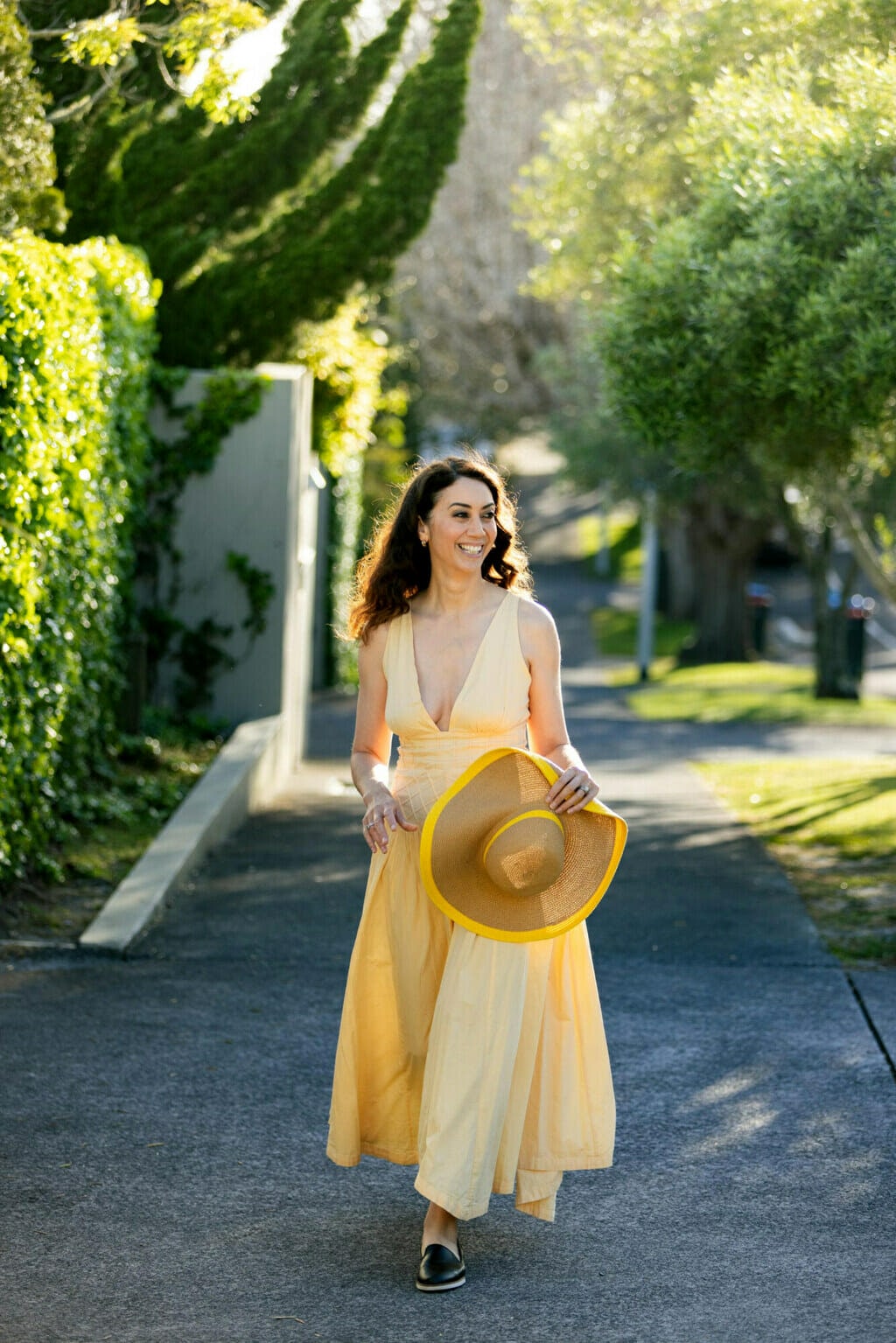 Miriama Kami walking down the street smiling. Miriama Kamo is wearing a yellow dress and holding a yellow straw hat. 