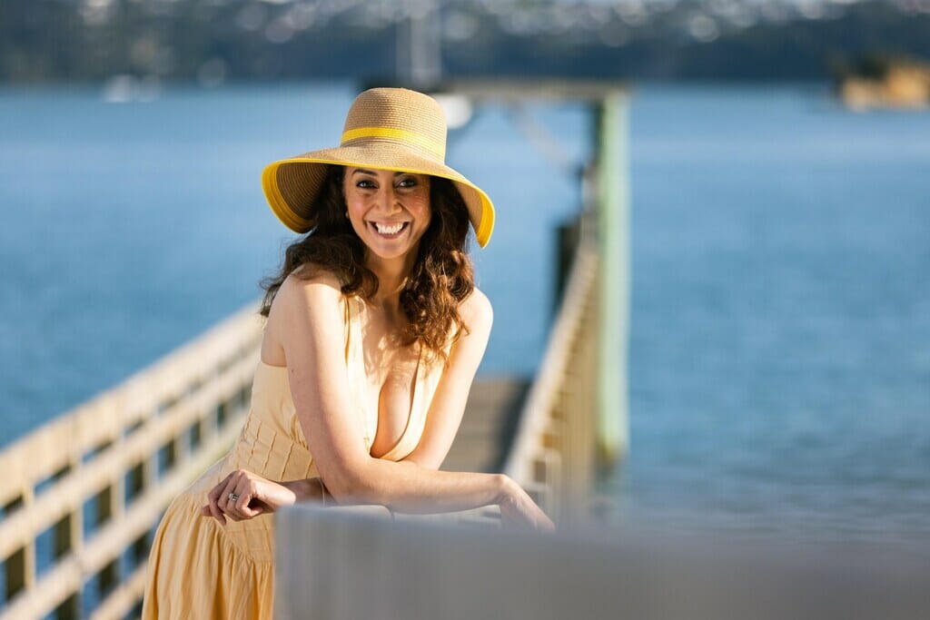 Miriama Kamo is posing on a wharf with water in the back. Miriama Kamo is wearing a yellow dress and a yellow straw hat.