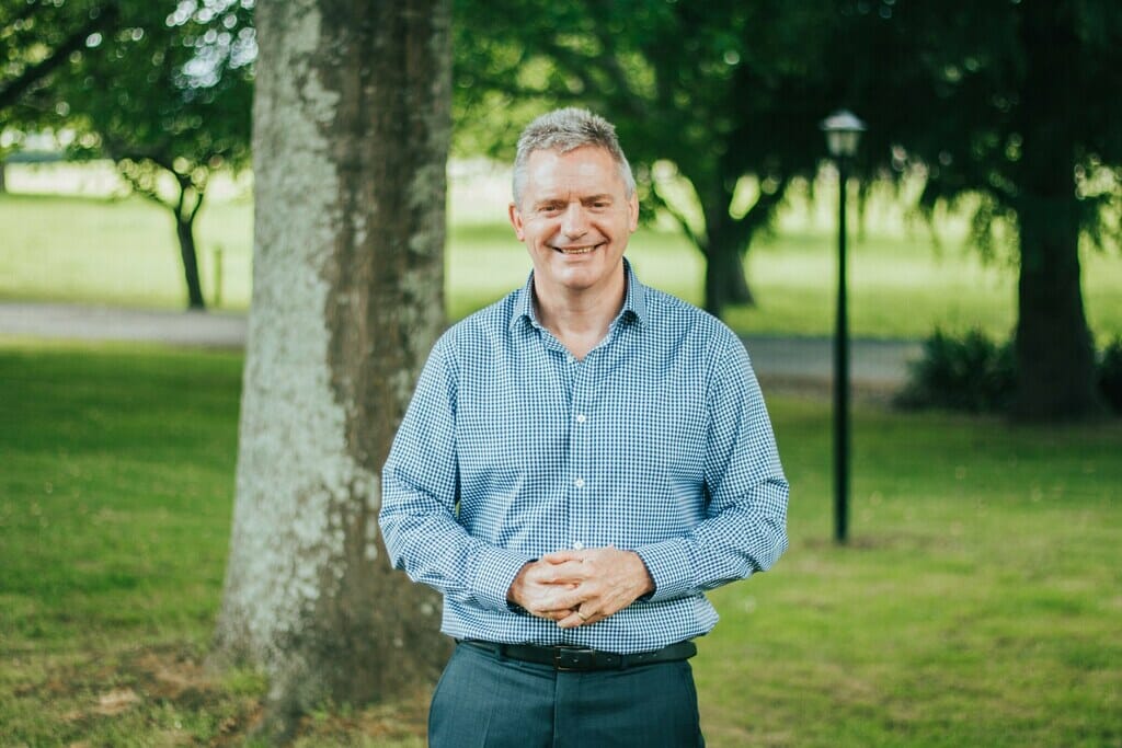 Mark Powell standing in front of tree holding hands together. 