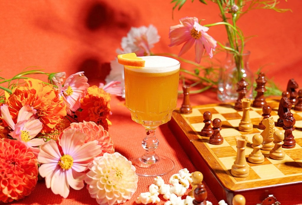 Orange rum cocktail with chessboard and flowers in front of orange backdrop