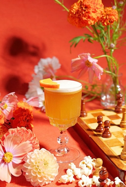 orange rum cocktail with chessboard and flowers in front of orange backdrop