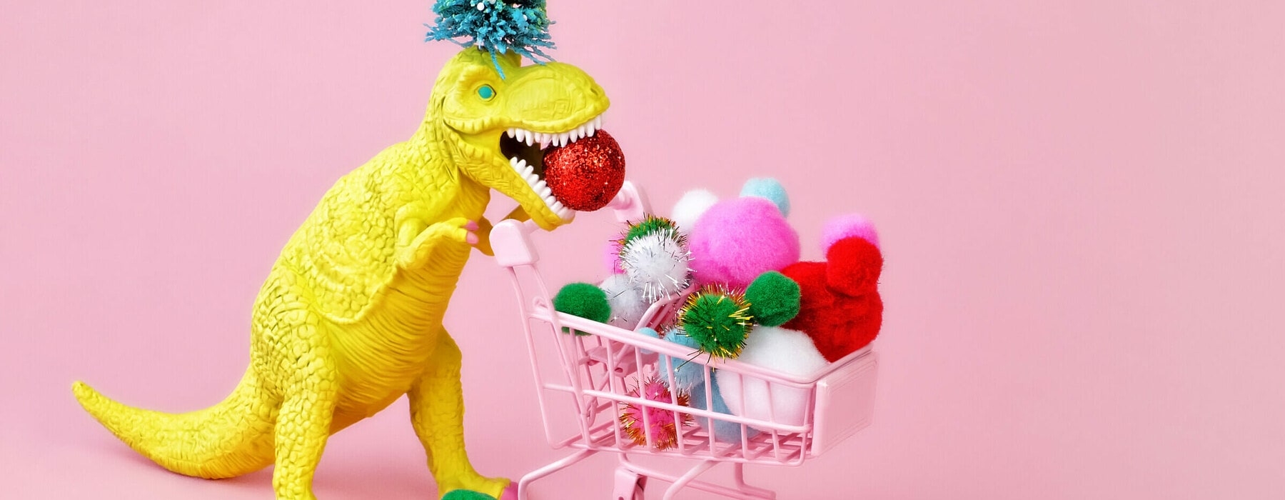 Yellow toy Tyrannosaurus rex wearing a Christmas tree on his head and filling a shopping cart with multicolored puff balls on pink background.