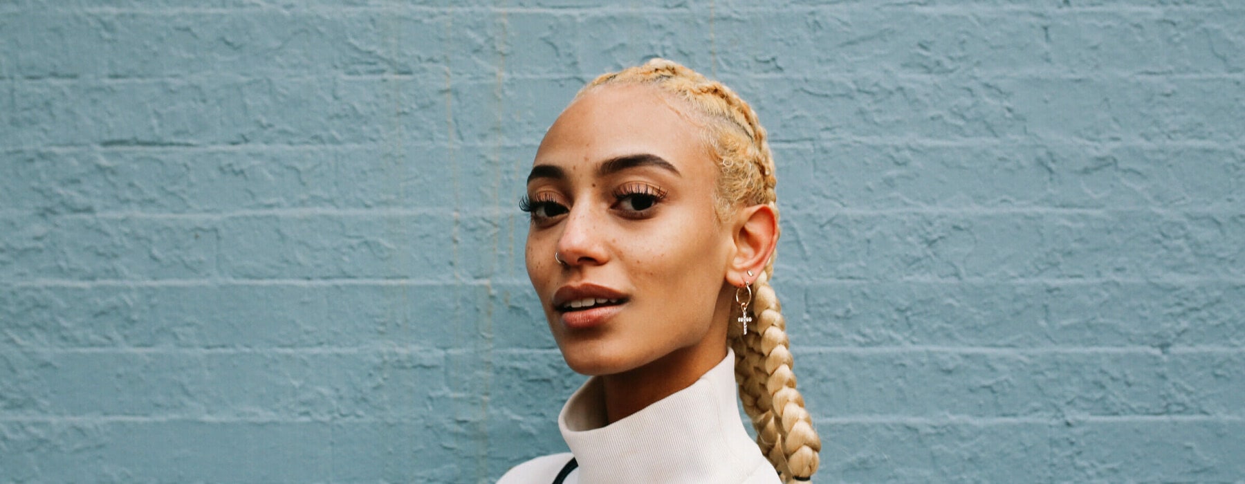 Vintage toned portrait of a young mixed race Latina woman from New York. She is sporting striking blond braids, leaning against the pastel toned brick wall.