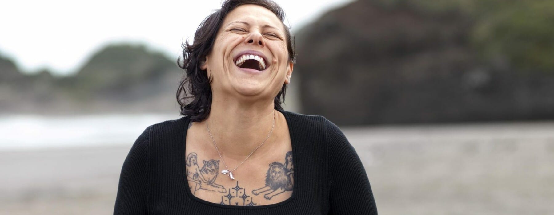 Anika Moa wearing black top and jeans standing at a beach and laughing