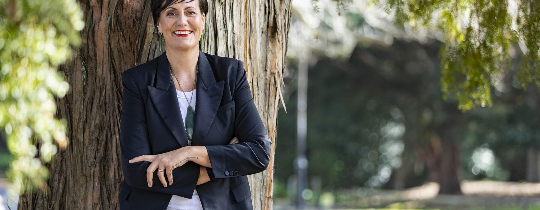 Rachel Taulelei leaning against a tree wearing a black suit and greenstone necklace