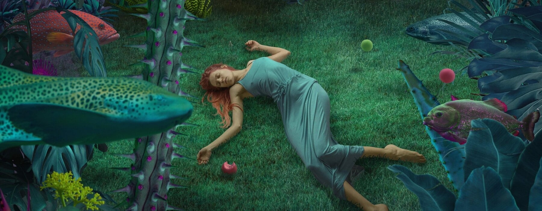 Woman with red hair and green dress lying on grass underwater with fish swimming by