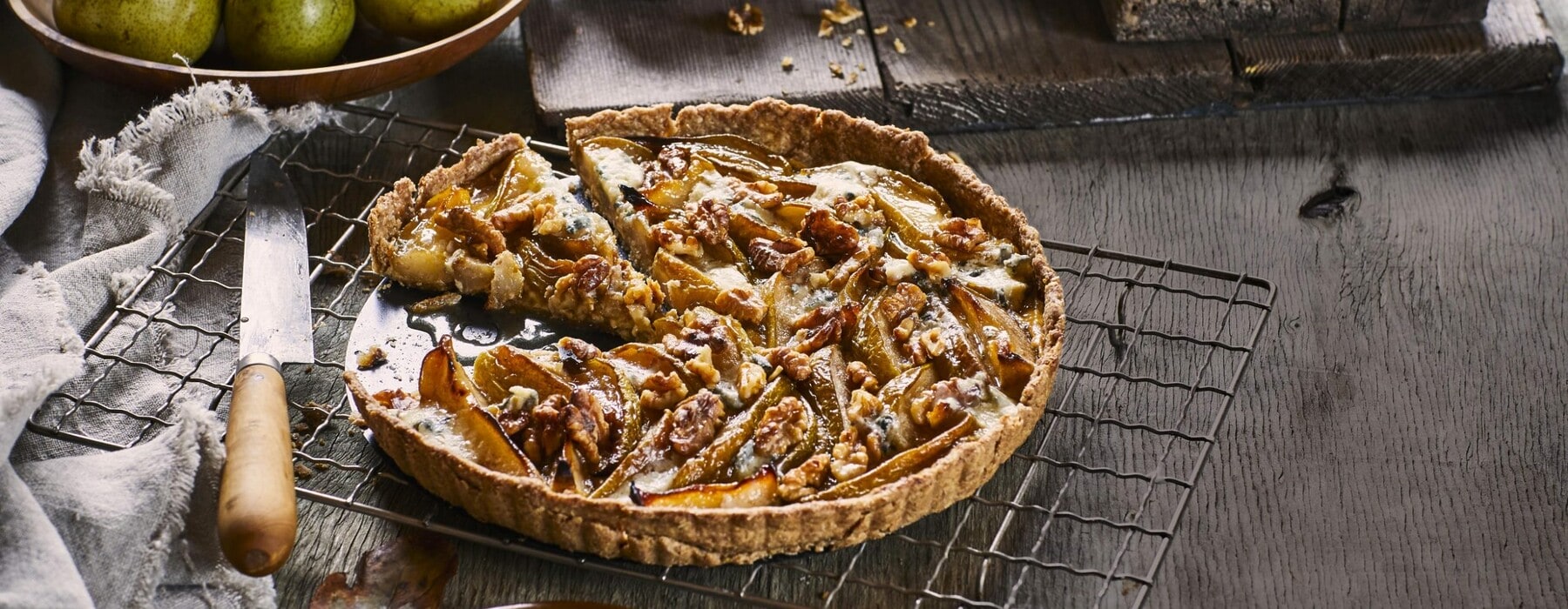 Pear, blue cheese and walnut tart being sliced by pair of hands while on a metallic oven rack