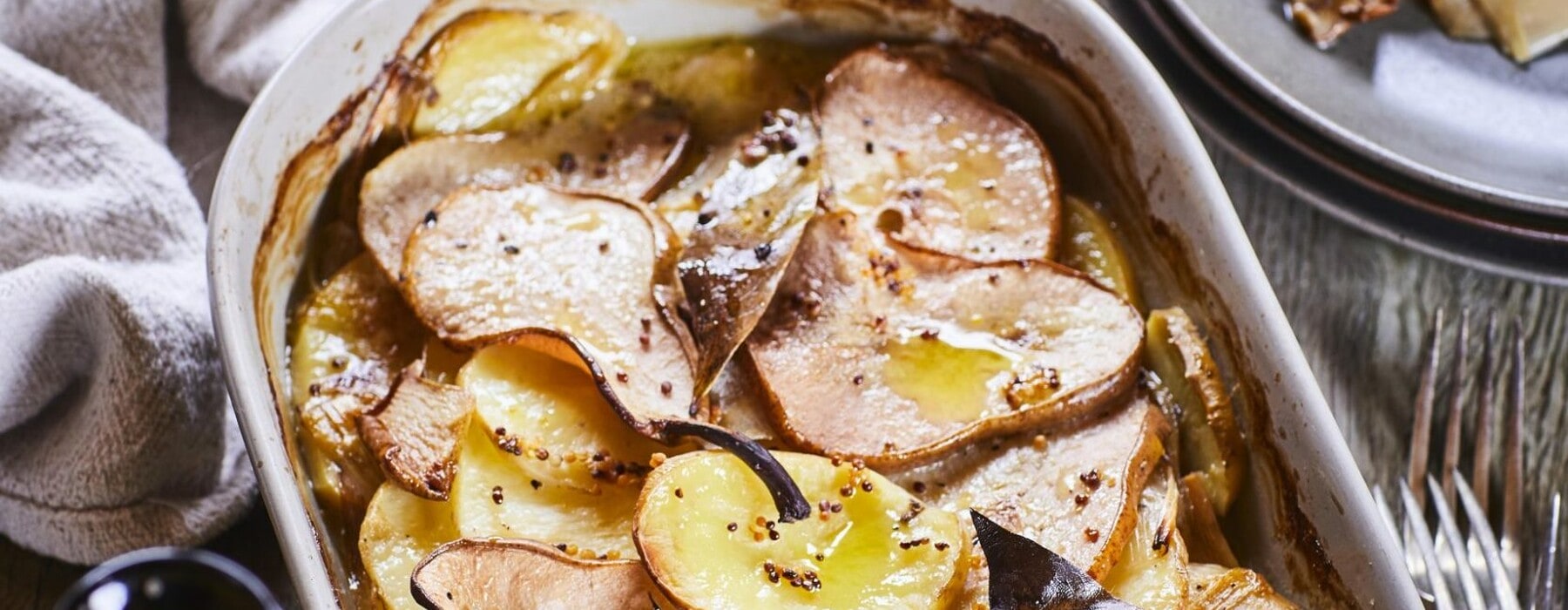 Potato, leek and pear bake served with cutlery on an oval shaped white dish set on top fo a brown table