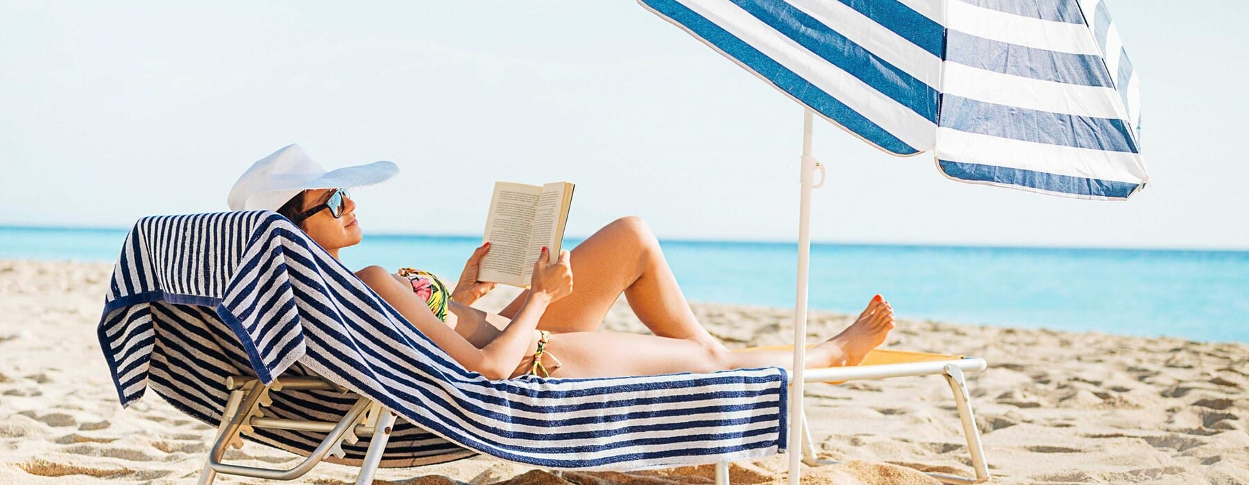 Woman reading on the beach under a blue striped umbrella on sun lounger